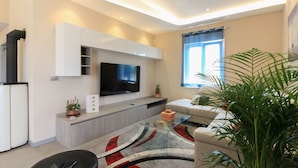 A modern living room with a flat-screen TV mounted on a white cabinet, a glass coffee table, a beige sofa, and a large potted plant.