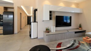 A modern living room with a flat-screen TV, built-in cabinets, a kitchen area with a refrigerator, and a dining area with a glass table.