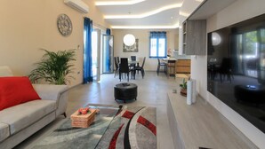 A modern living room with a sofa, a coffee table with fruit, a dining area with chairs, a kitchen with a TV, and a large window.