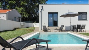 A modern outdoor pool area with a clear blue pool, black lounge chairs, a table with chairs, and a large umbrella. - Villa Relax (Istria - Sisan) (Sisan)