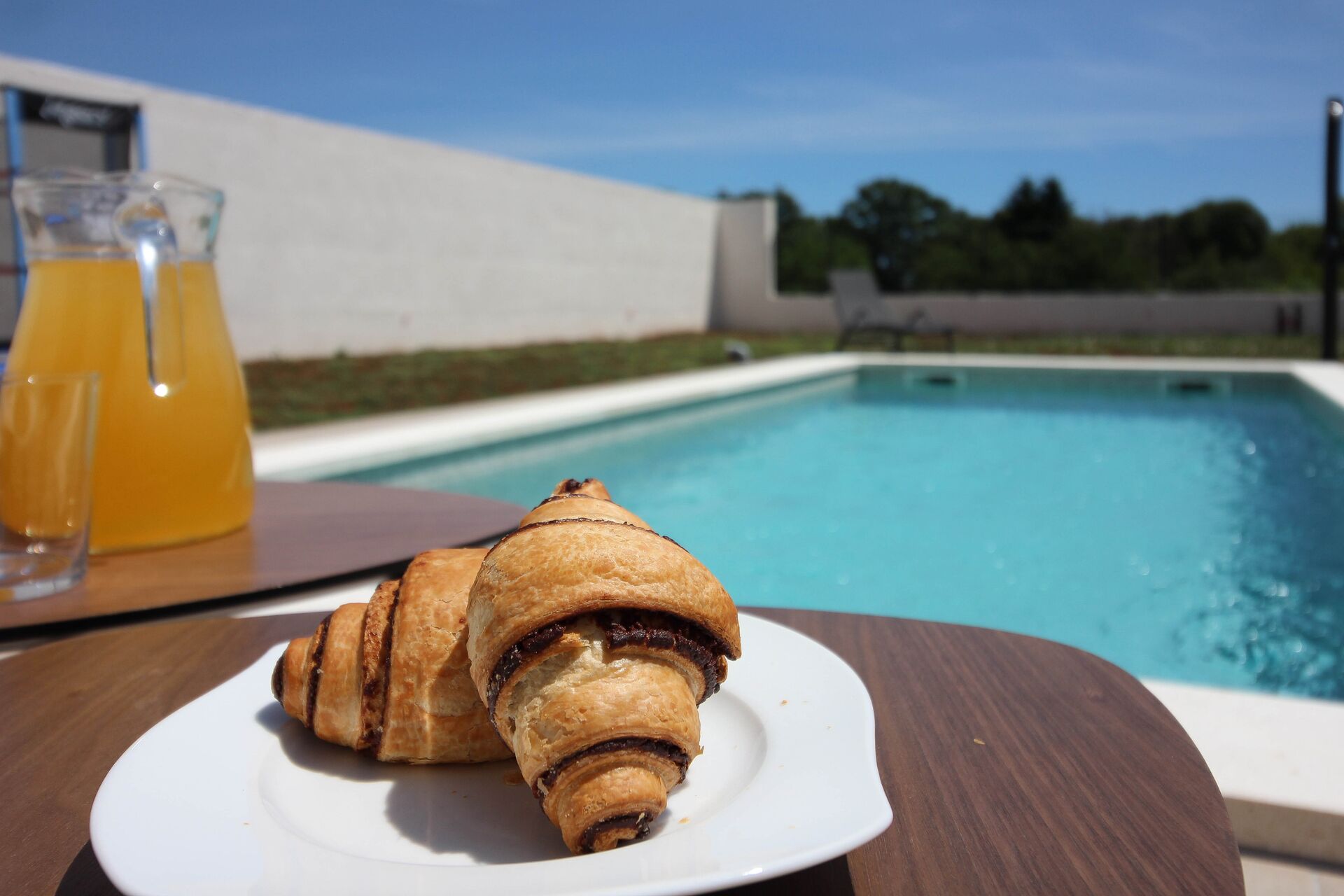 Un croissant sobre un plat blanc a la vora d'una piscina, amb un got de suc d'aranja.