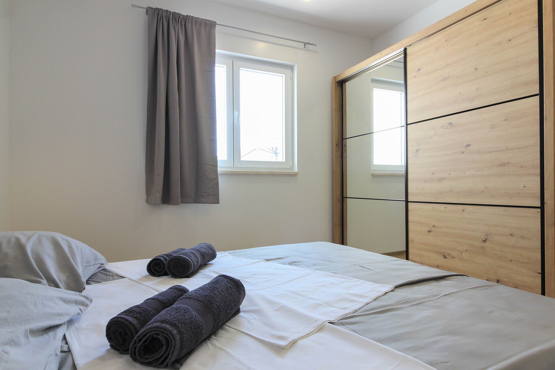 A bedroom with a bed, grey curtains, a window, and a wooden wardrobe.