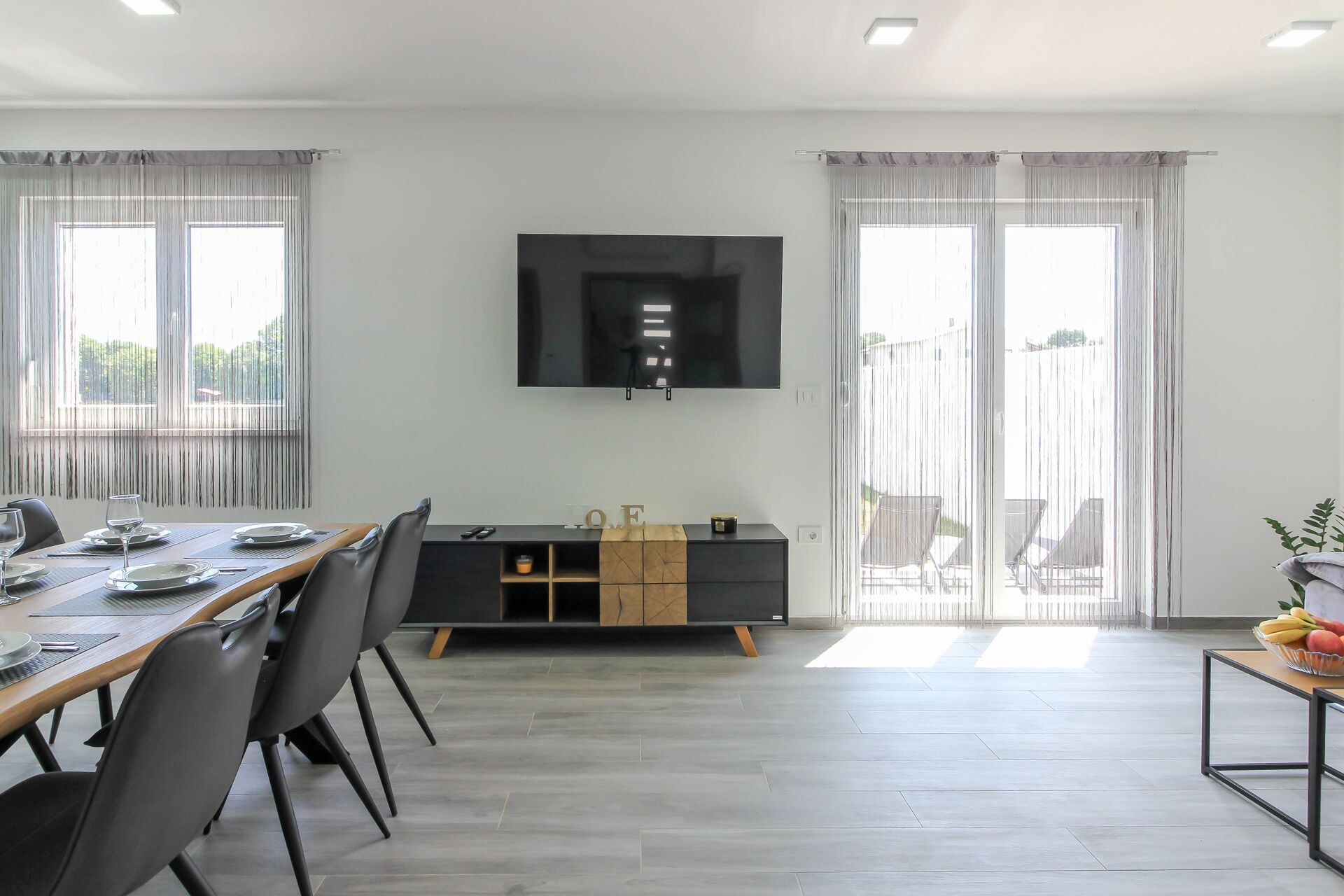 A modern living room with a dining area, a flat-screen TV mounted on the wall, and large windows with sheer curtains.