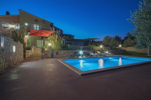A swimming pool with lounge chairs and an umbrella, surrounded by stone walls and a building with multiple windows.