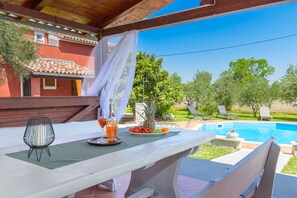 Un área de comedor al aire libre techada, con una mesa dispuesta para dos personas, una piscina al fondo y un edificio rojo con tejado de baldosas.