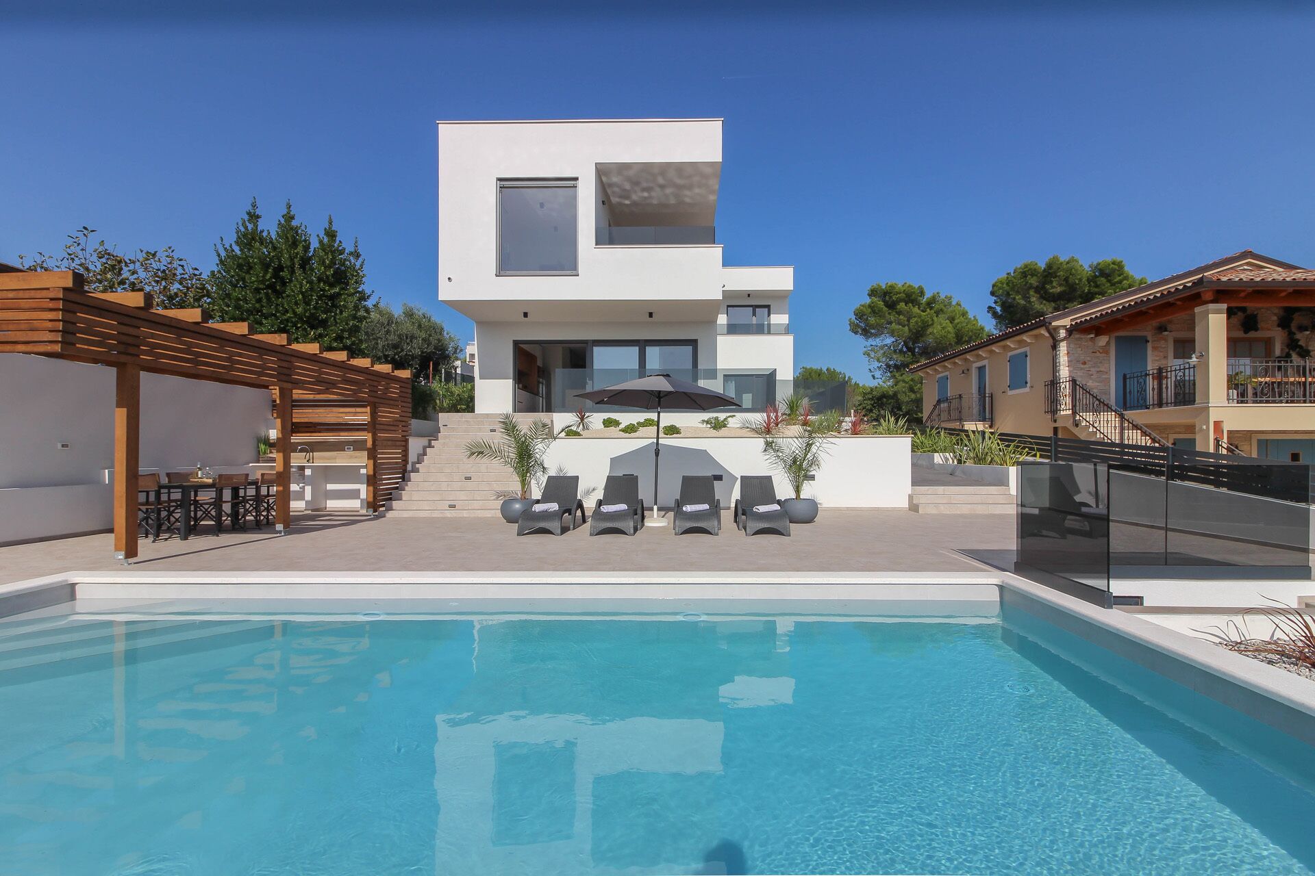 A modern poolside area with a clear blue pool, a wooden pergola, and a two-story building with a balcony.