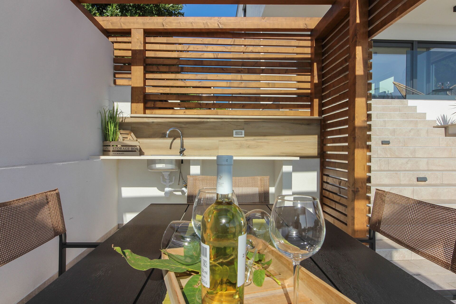 Een houten pergola met een ingebouwde wasbak en een opbergkist, een tafel met wijnglazen en een uitzicht op een modern gebouw op de achtergrond.