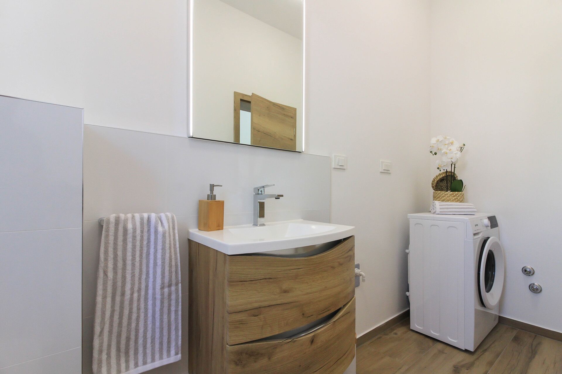 A modern bathroom with a wooden vanity, a white sink, a mirror, a towel, a washing machine, and a small plant.