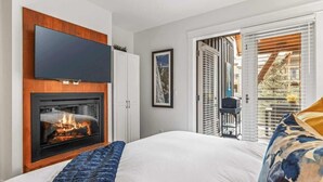 A bedroom with a fireplace, a flat-screen TV mounted above it, a bed with a blue and white patterned blanket, and a view of the outside through a door with blinds. - Vista Peak Hydeaway at Solara Resort (Canmore)