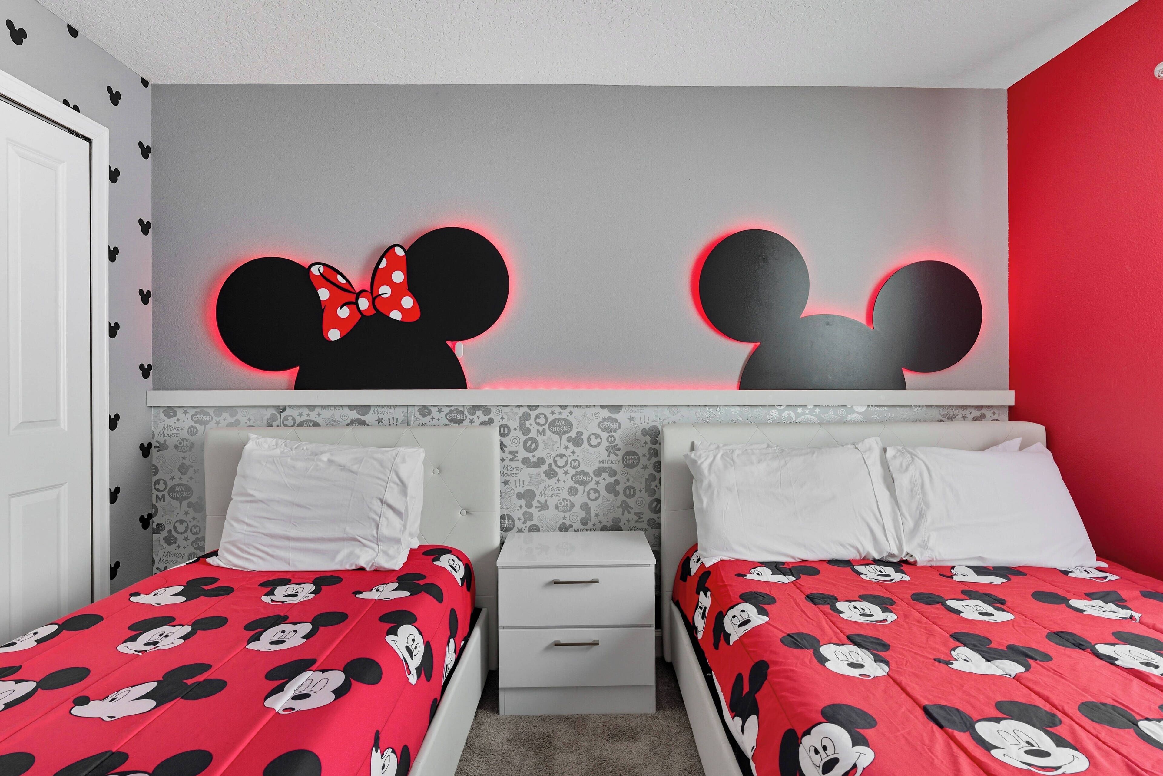 Un dormitorio con dos camas, cada una con ropa de cama temática de Mickey Mouse. Hay dos mesitas de noche con cajones, y las paredes están decoradas con motivos de Mickey Mouse.