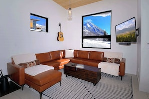 A modern living room with a sectional sofa, a coffee table, a guitar, and three large windows with mountain views.