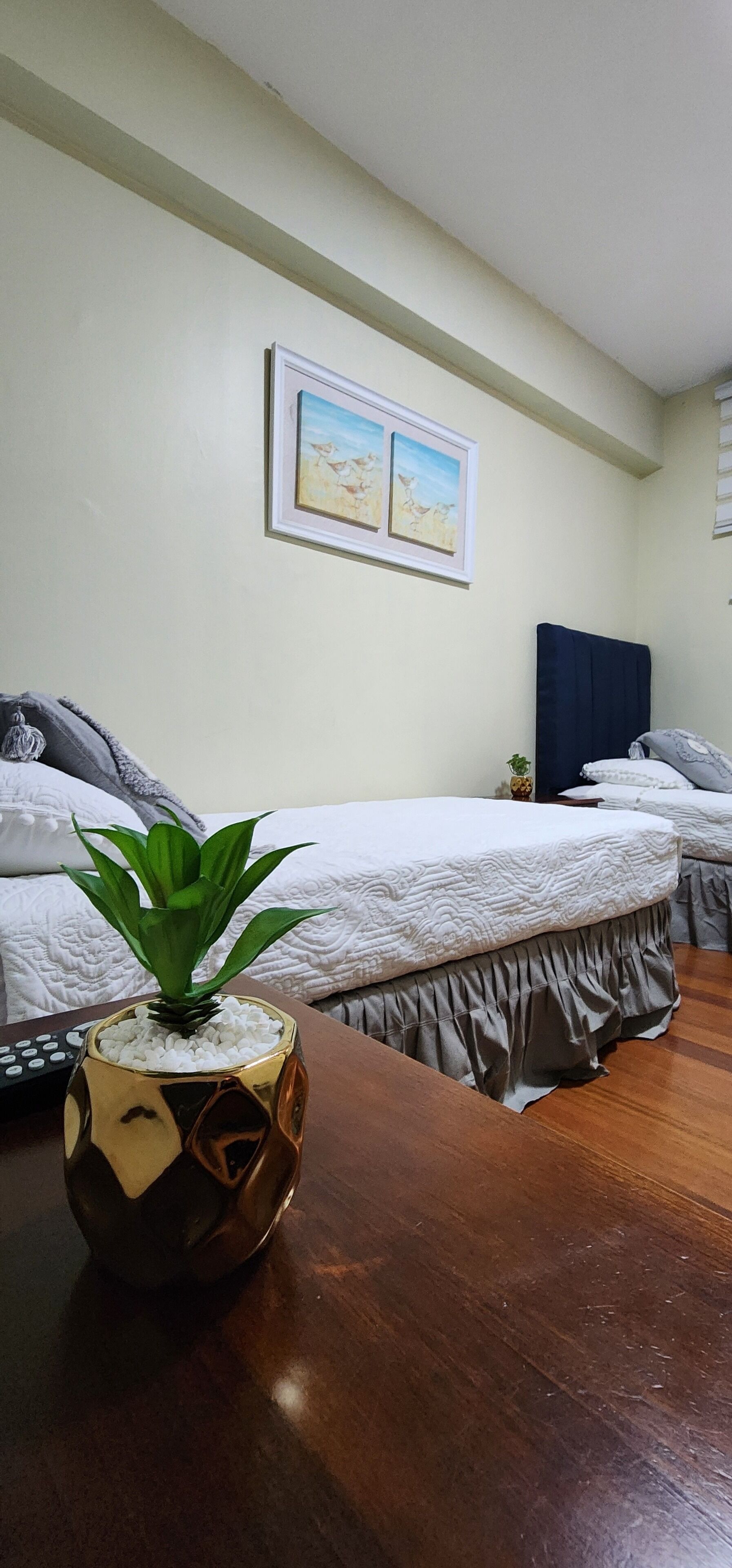 A hotel room with two beds, a wooden table, a potted plant, a painting on the wall, and a remote control on the table.
