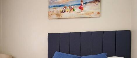 A neatly made bed with blue and white bedding, a painting of a beach scene on the wall, and a lamp on the side.