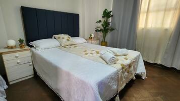 A neatly made bed with a white duvet and decorative pillows, a bedside table with a plant and a small lamp, and a large window with curtains.