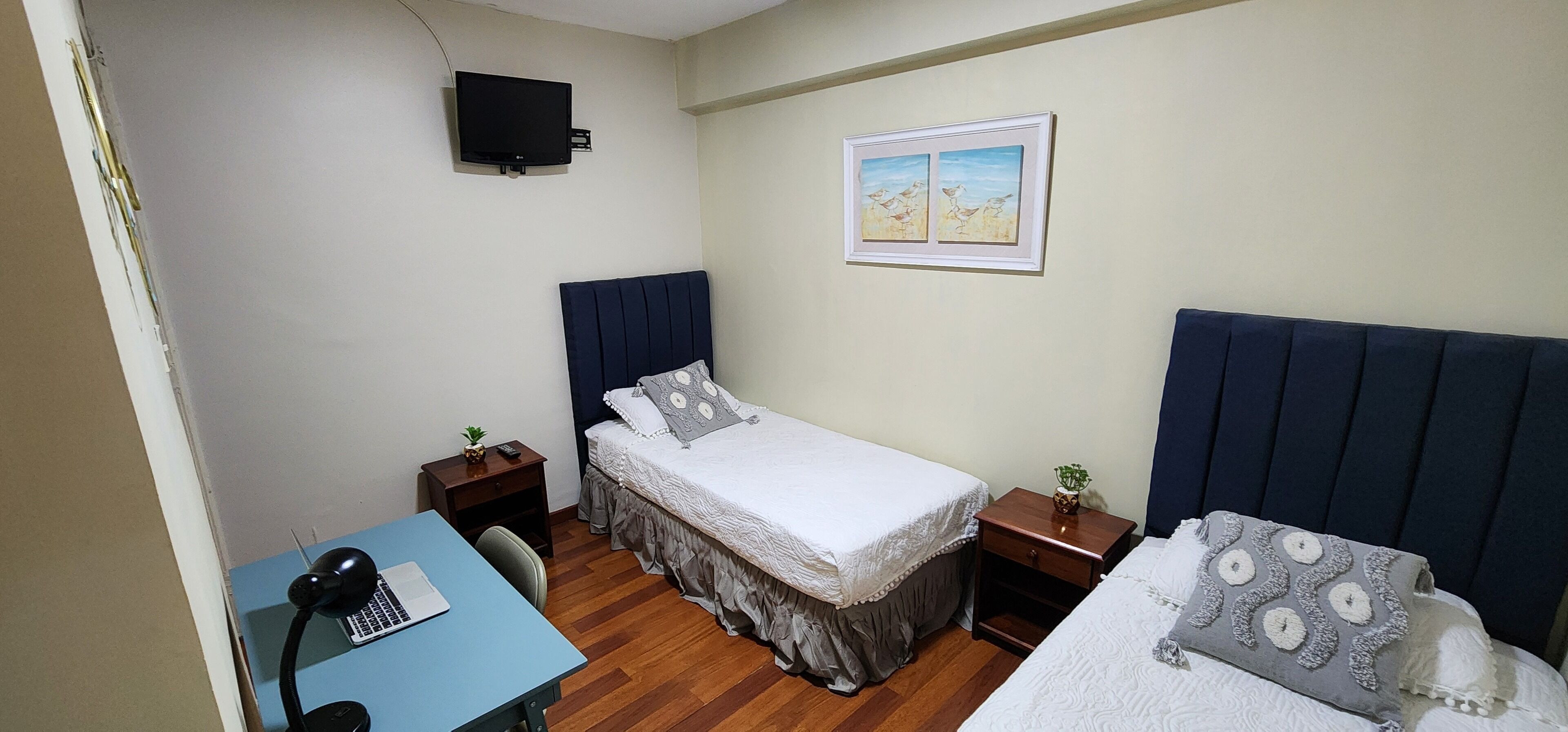 Quarto pequeno e arrumado com uma cama de solteiro, uma escrivaninha com um laptop, uma TV fixada na parede e um quadro emoldurado sobre a cama.
