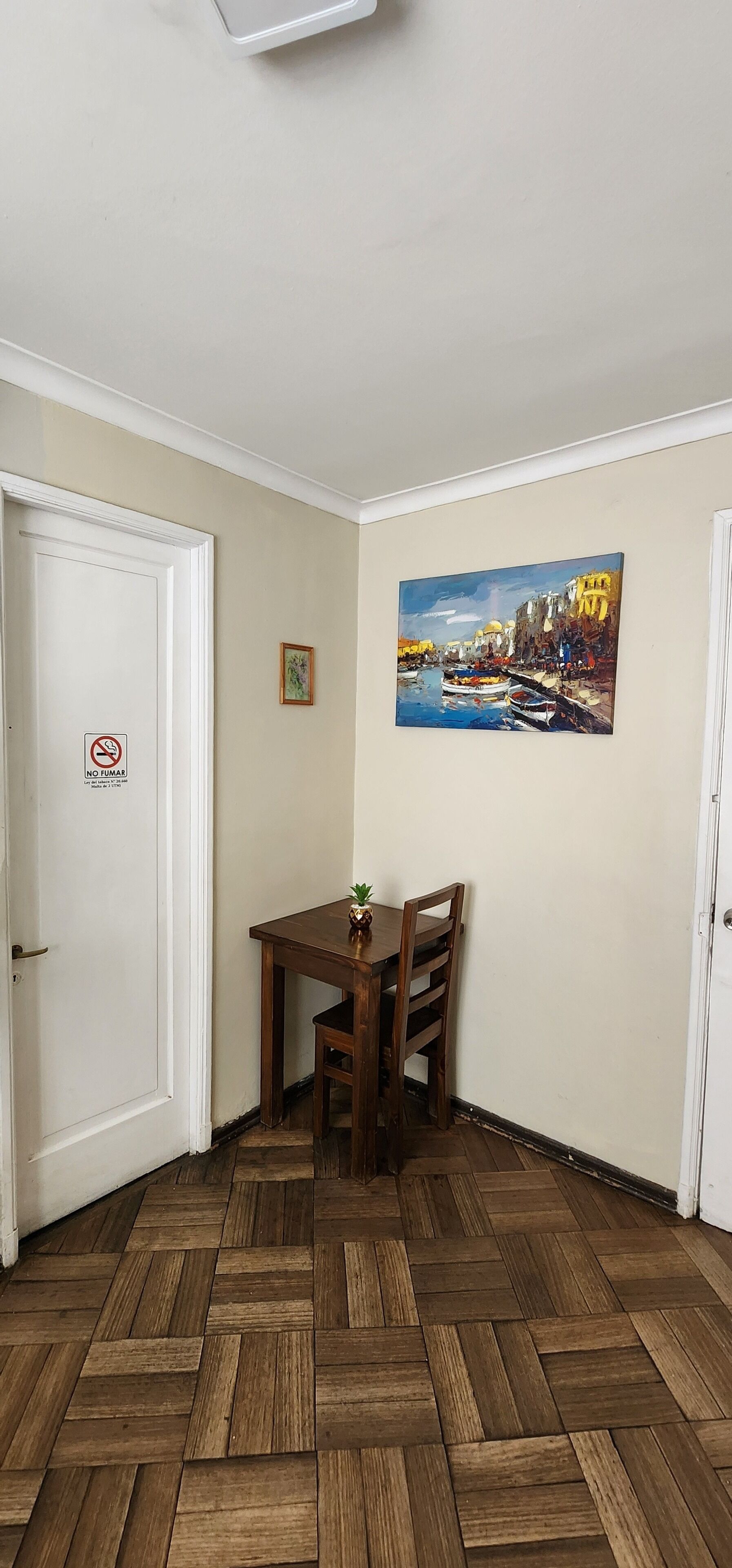 A corner room with a wooden herringbone floor, a dining table with chairs, a painting on the wall, and a door with a 'NO TUMBLING' sign.