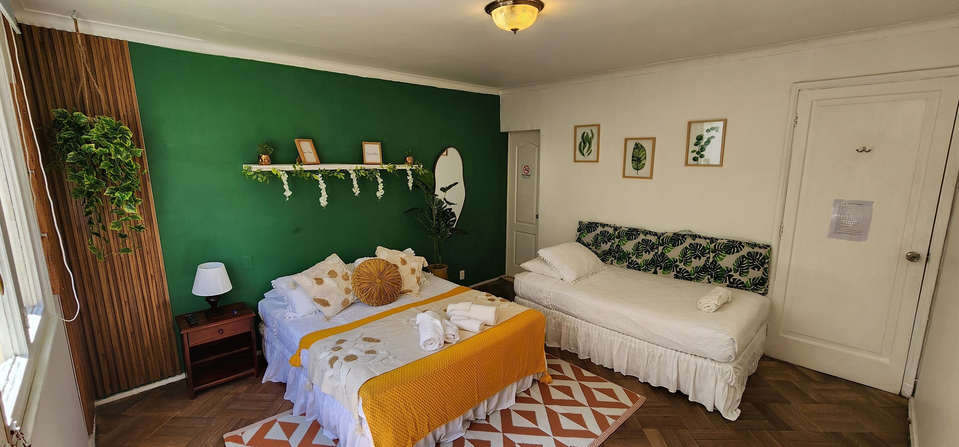 A room with a bed, a sofa, a green wall, a wooden slat wall, a mirror, and a shelf with framed pictures and plants.