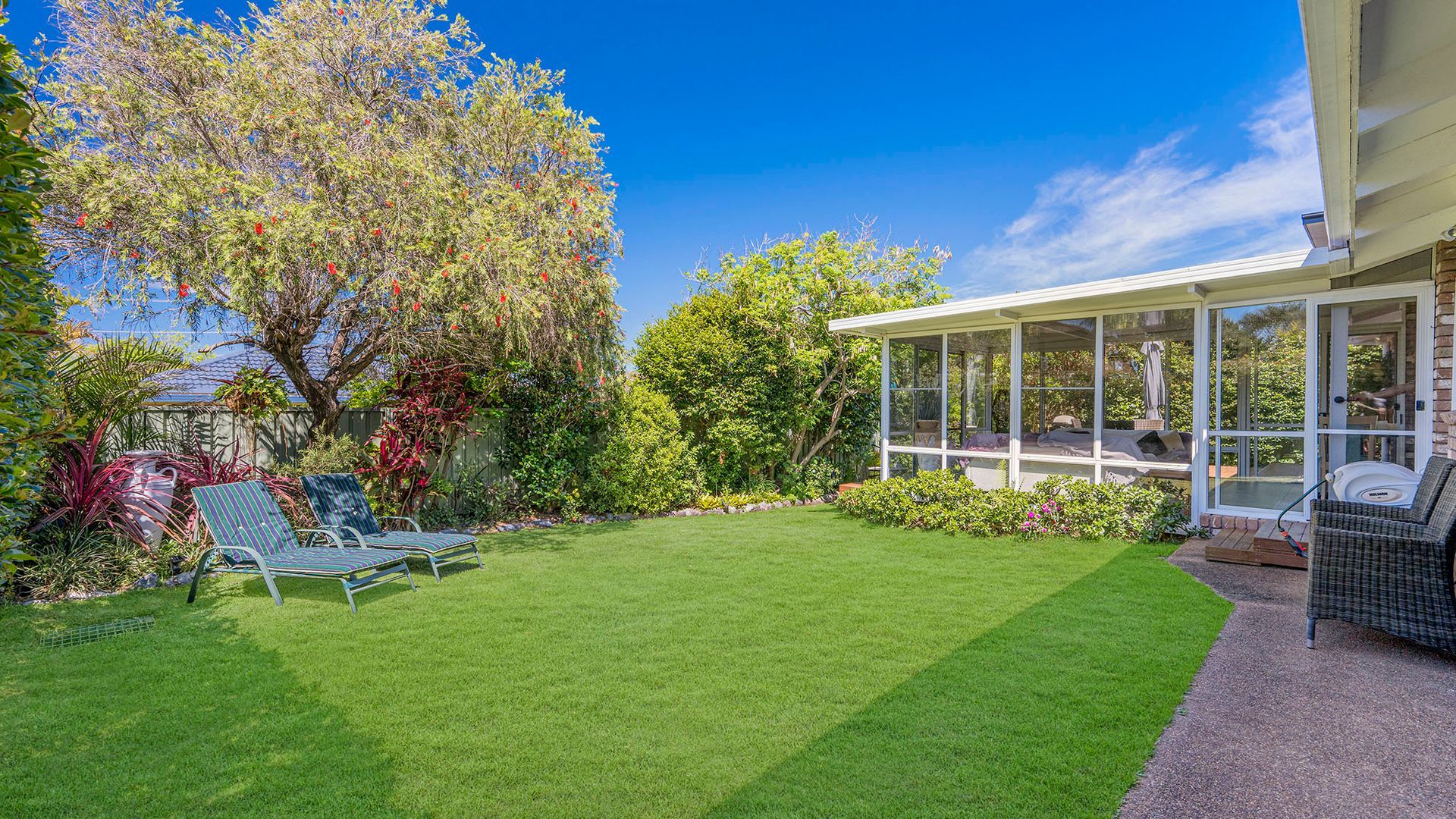 A well-maintained backyard with a lawn, two lounge chairs, a covered patio area, and a variety of plants and trees.