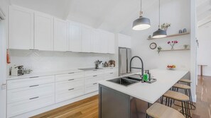 A modern kitchen with white cabinetry, a stainless steel refrigerator, a sink with a faucet, and a bar area with stools. - Lighthouse Beach Luxury (Port Macquarie)