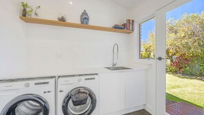 A compact laundry area with two washing machines, a sink, and a window overlooking a garden. - Lighthouse Beach Luxury (Port Macquarie)