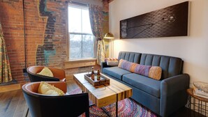 A cozy living room with a brick wall, a large window with curtains, a blue sofa, two black chairs, a wooden coffee table, and a modern art piece. - BRAND NEW! Vibrant & Exquisite -1 Mile to Downtown (Nashville)