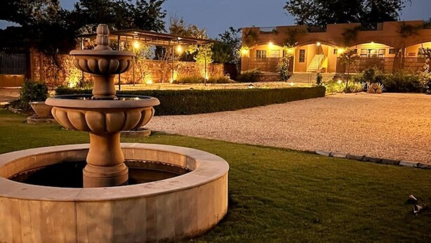 Un espacio exterior bien iluminado con una fuente de piedra, un seto cuidado y un edificio con varias ventanas y una escalera.