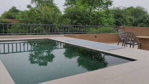 Una piscina con plataforma de baldosas, rodeada por una barandilla metálica. Hay dos sillas de mimbre y una pequeña mesa en la plataforma.