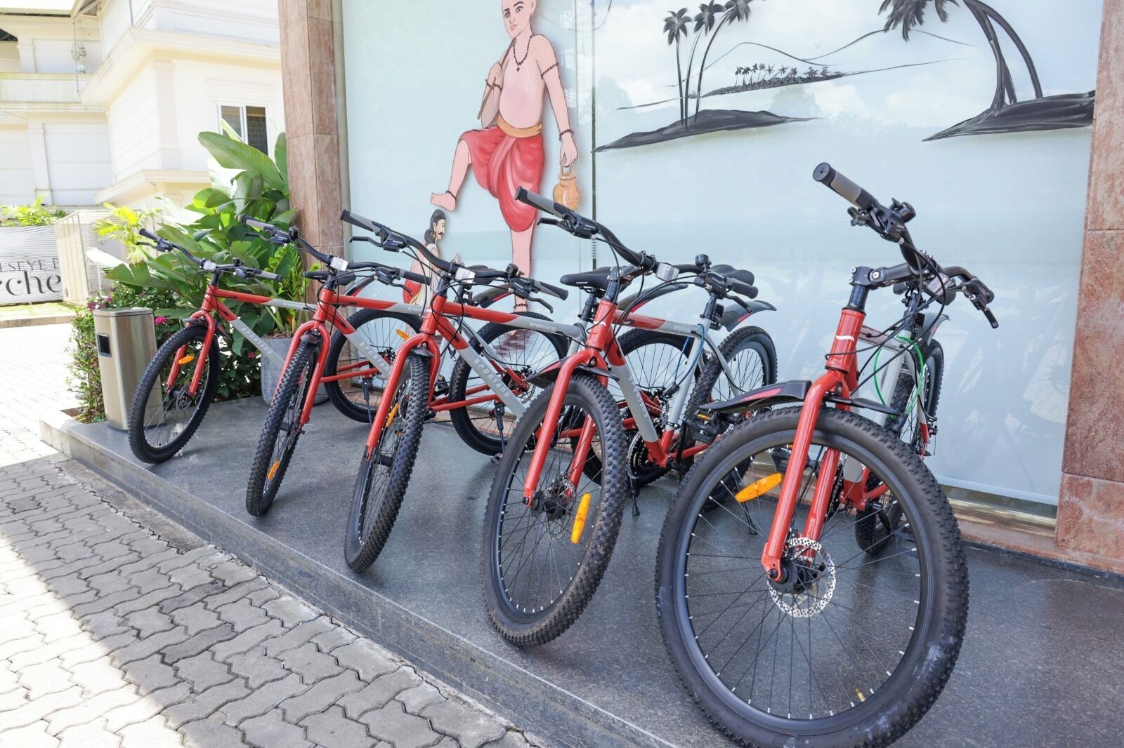 A row of red bicycles parked outside a building with a mural of a person and palm trees.