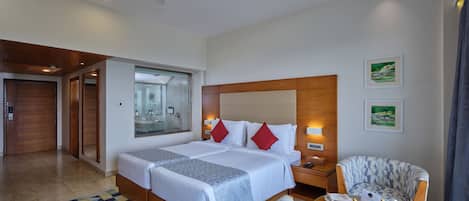 Habitación de hotel con una cama grande, ventilador de techo, ducha de vidrio, cabecero de madera, una pequeña mesa con fruta y un sillón estampado.