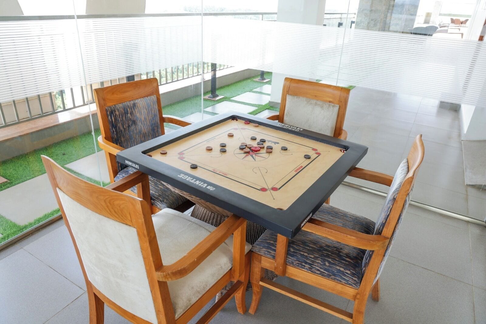 Una mesa de fútbol de mesa con cuatro sillas, ubicada en una habitación luminosa con ventanas grandes y vistas a un área verde al aire libre.