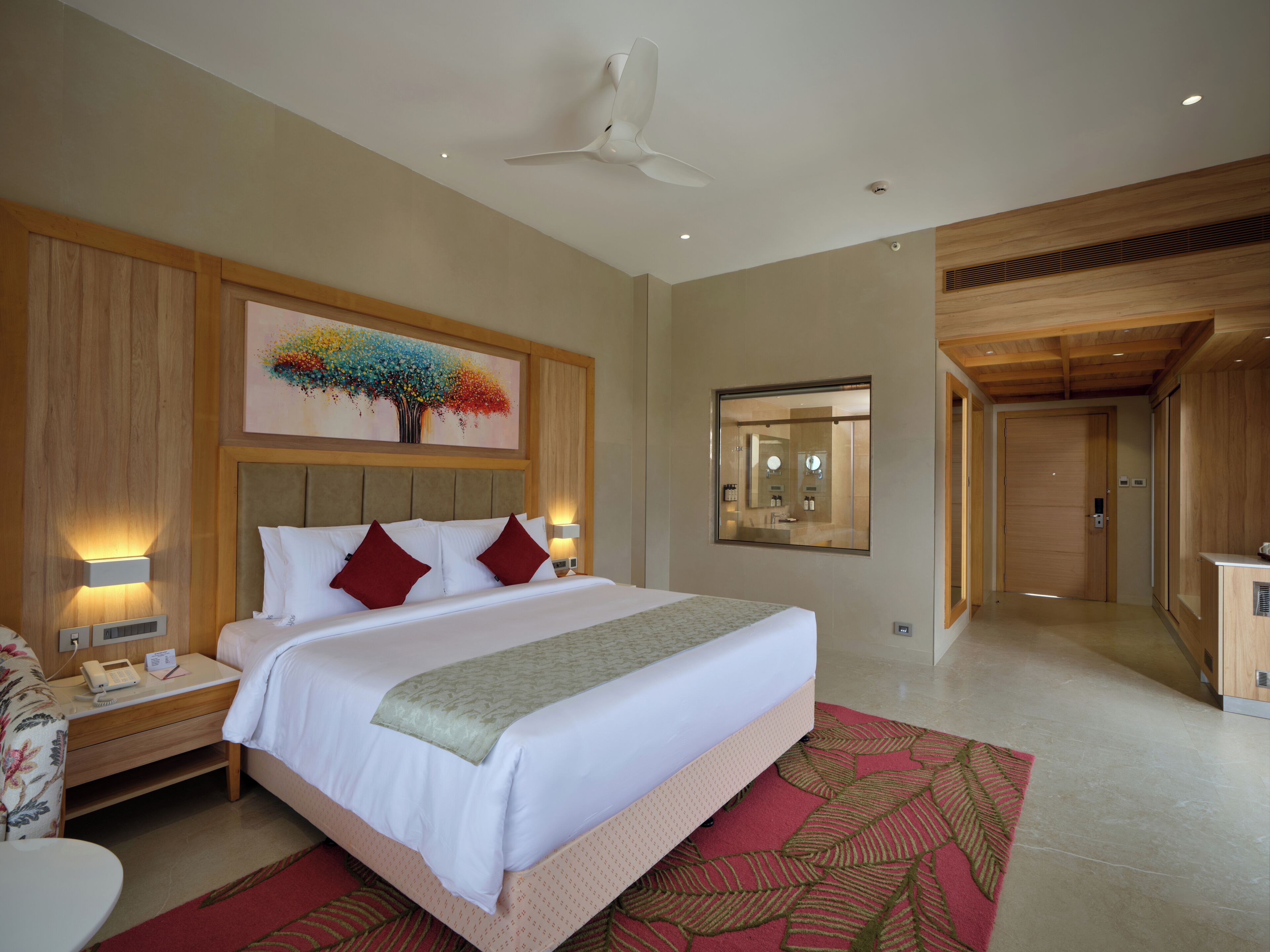 A modern hotel room with a large bed, a ceiling fan, a painting on the wall, and a bathroom mirror.