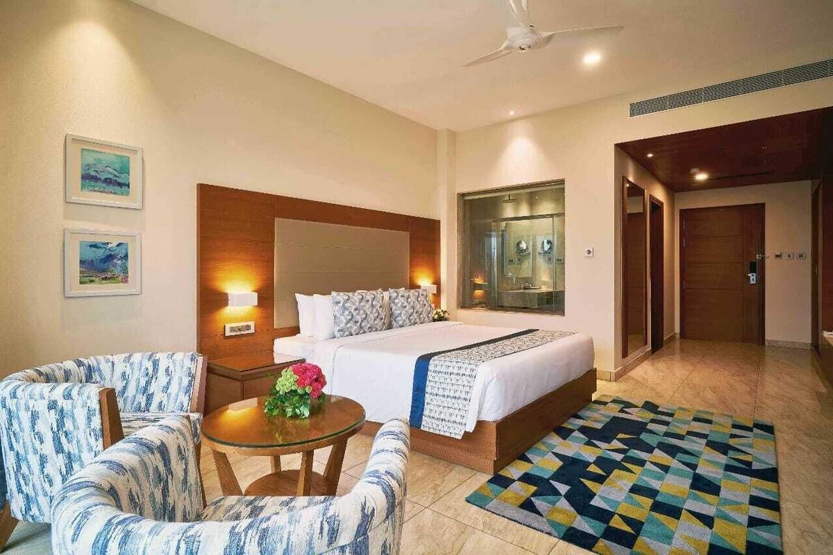 Ein Hotelzimmer mit einem Bett, zwei Sesseln, einem kleinen Tisch mit Blumen, einem gemusterten Teppich und einem Deckenventilator.