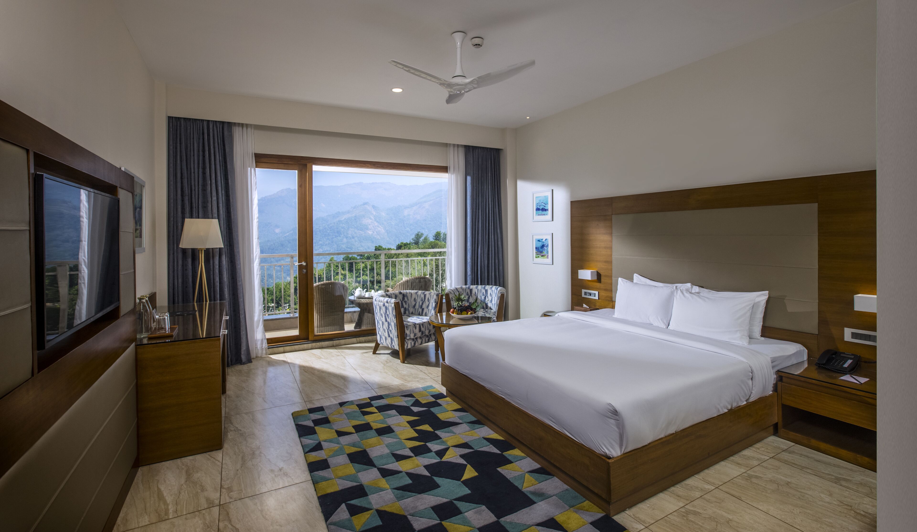 Een hotelkamer met een groot bed, een televisie, een balkon met uitzicht op bergen en een plafondventilator.
