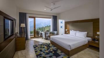 A hotel room with a large bed, a TV, a balcony with a view of mountains, and a ceiling fan.