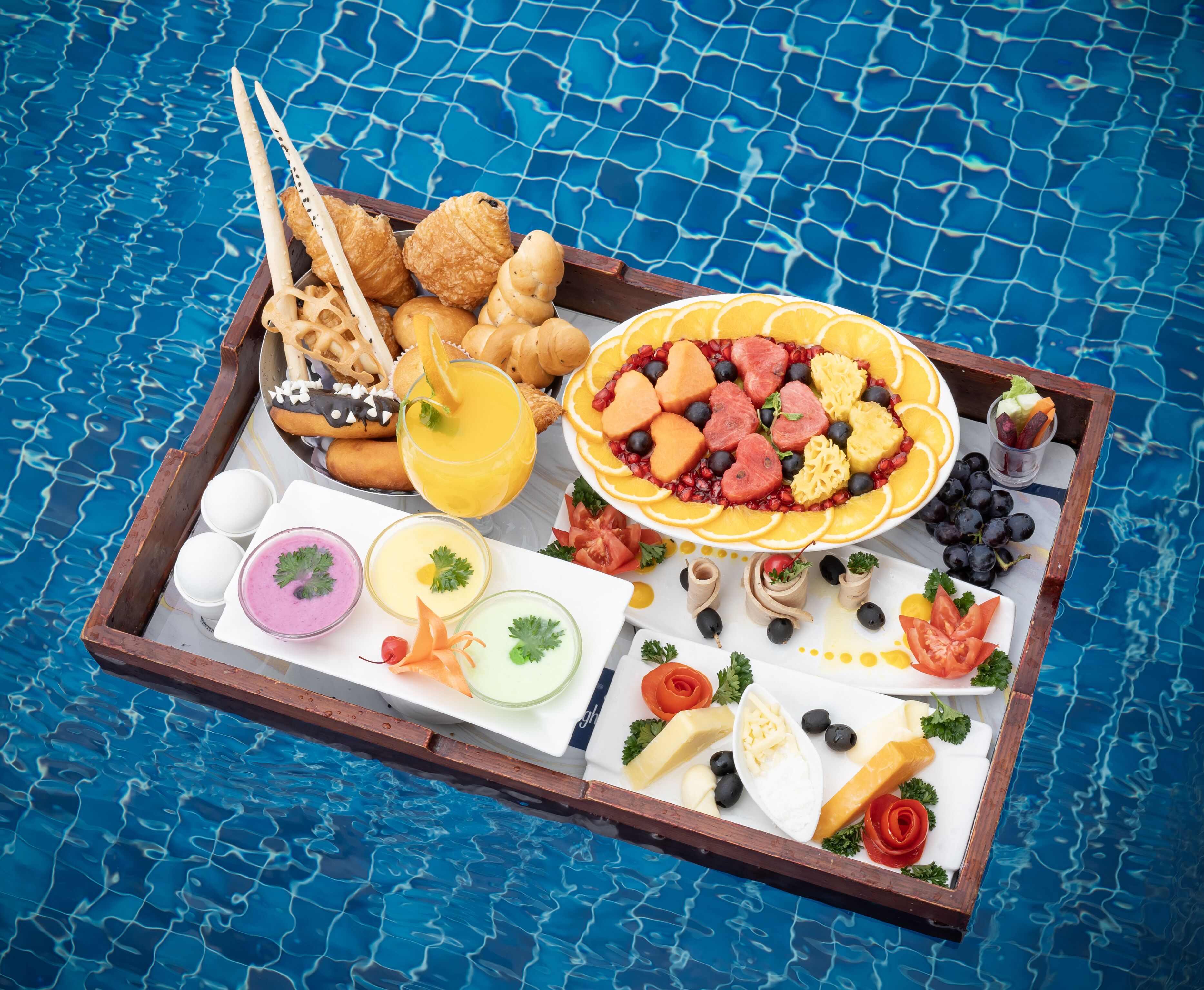 A tray of food items including croissants, fruit, cheese, and olives, placed on a wooden board by a pool.