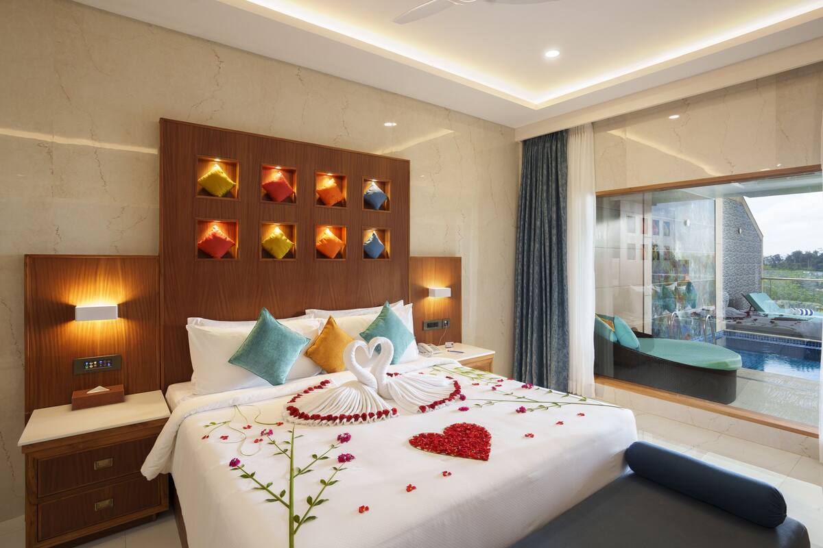 Ein Hotelzimmer mit einem großen Bett, einem Holzkopfende, einem Nachttisch mit Lampe und Blick auf einen Pool und Liegestühle durch eine Glastür.