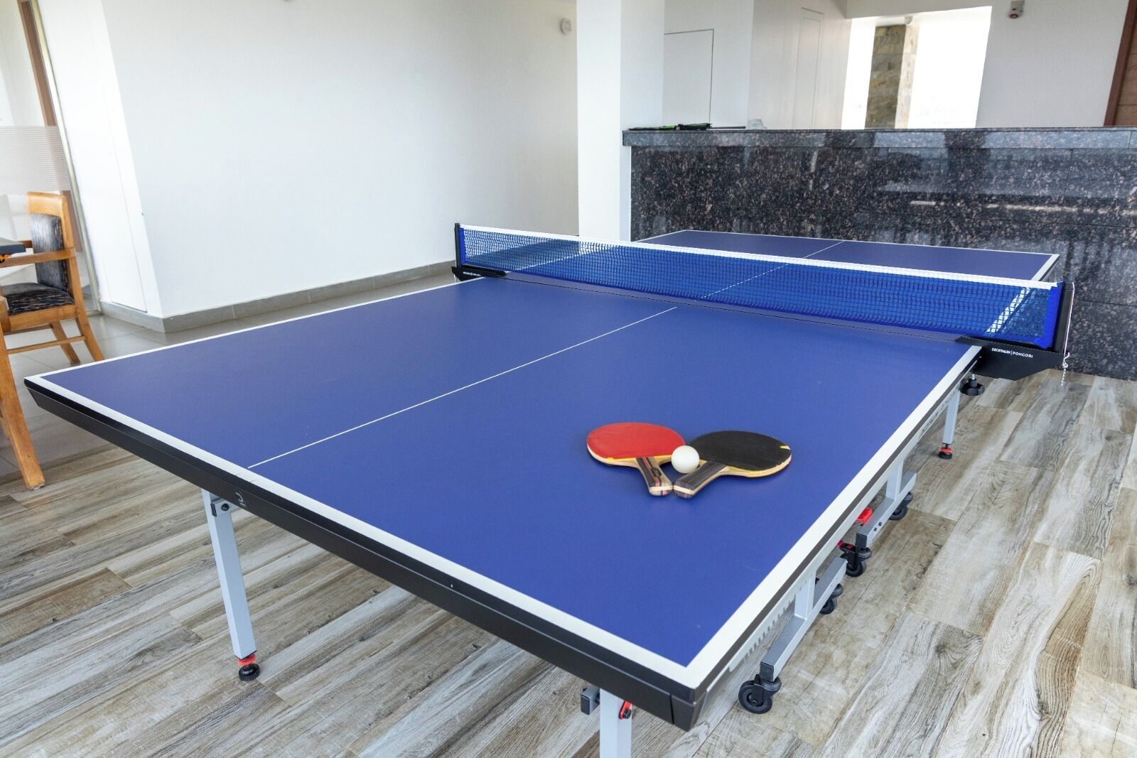 A ping pong table with two paddles and a ball in a room with wooden flooring and a stone wall.