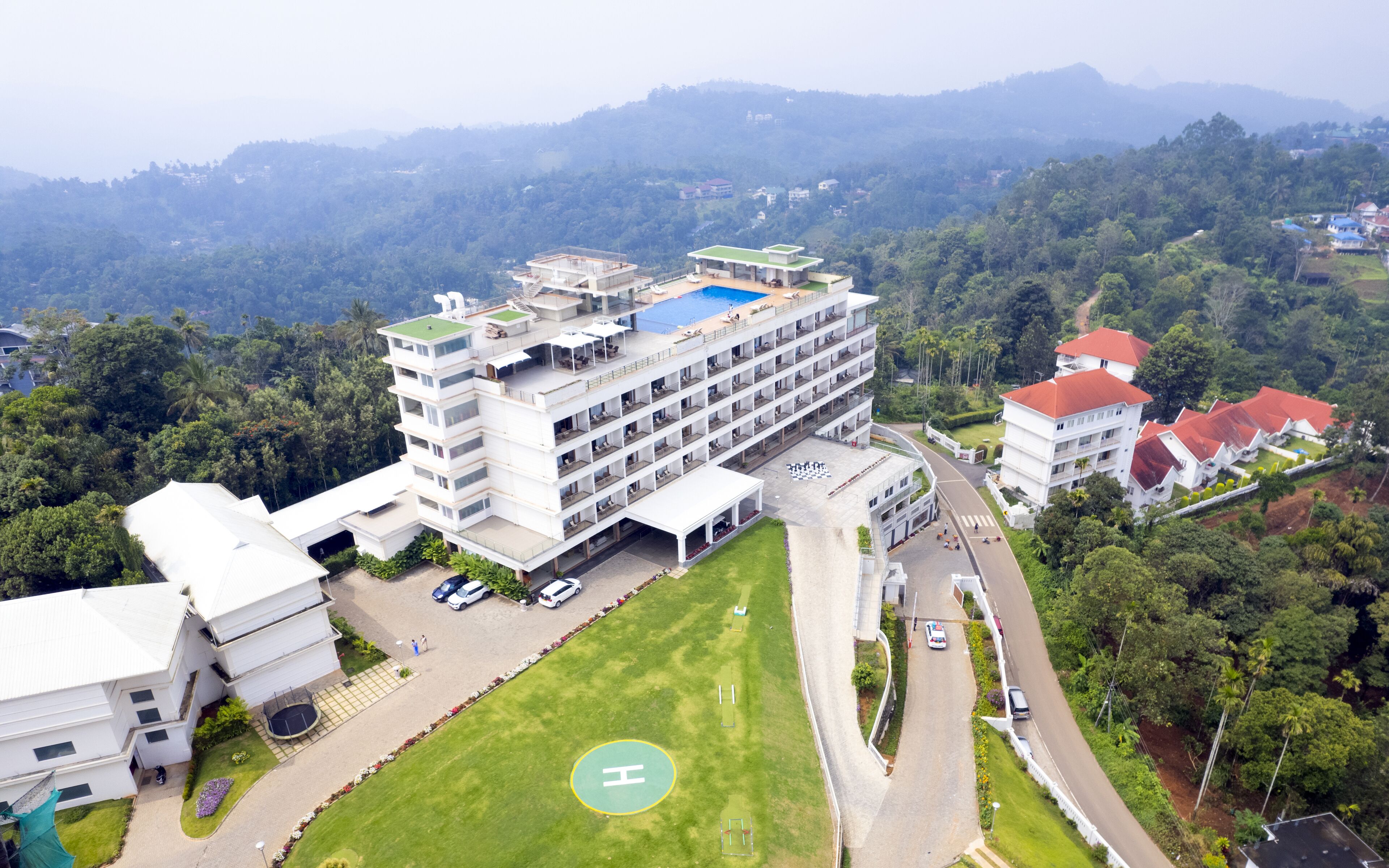 A multi-story hotel with a rooftop pool, surrounded by lush greenery and hilly terrain.