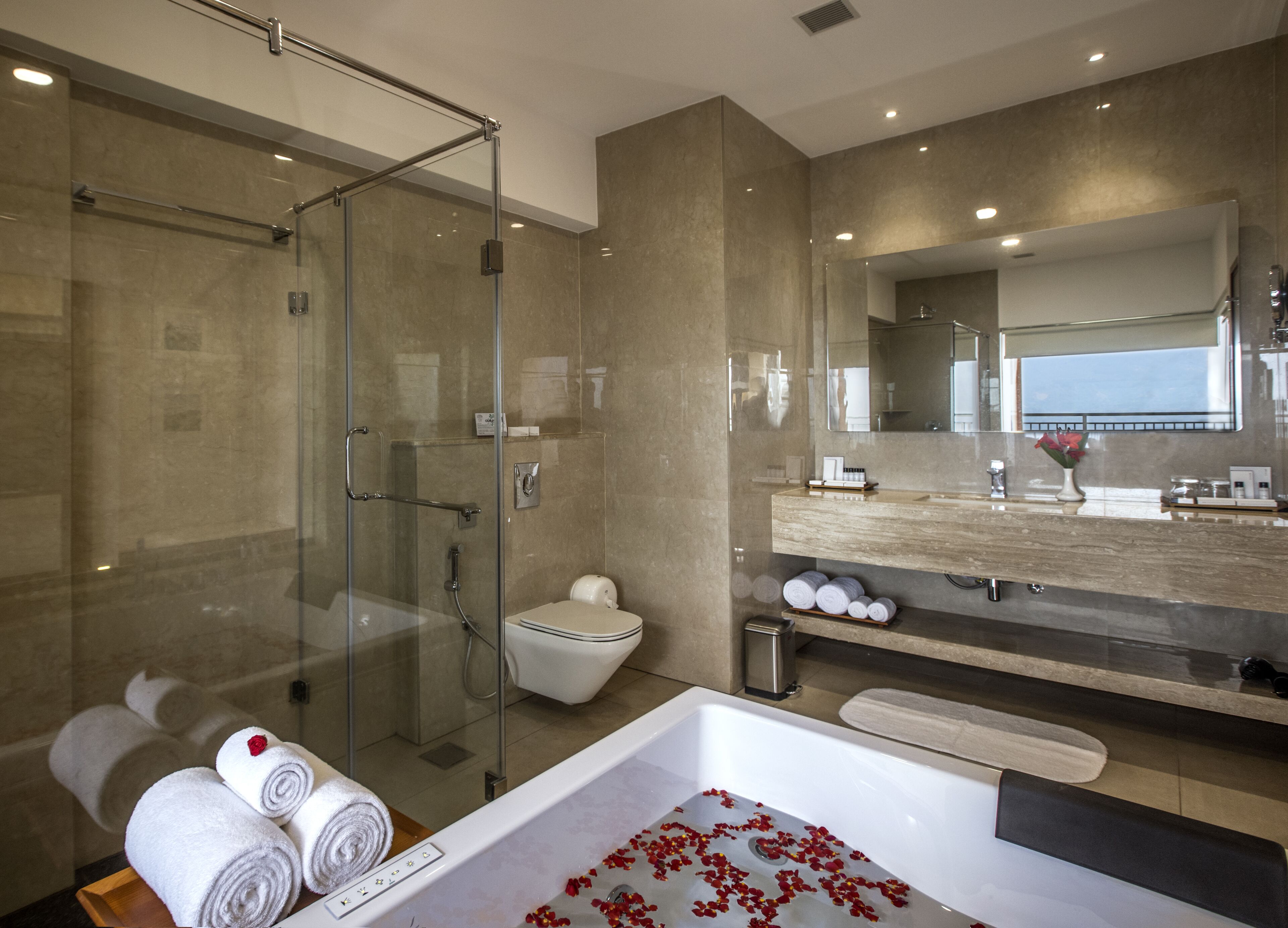 Un baño moderno con ducha de vidrio, inodoro blanco, una bañera grande con pétalos de rosa y un tocador con espejo.