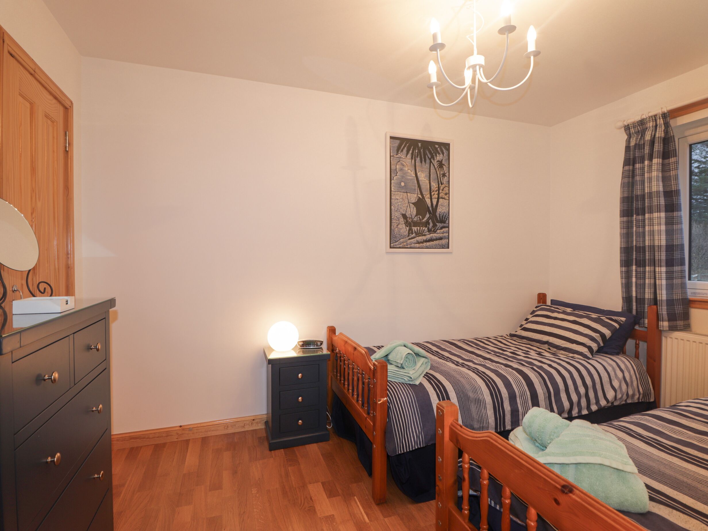 A bedroom with a wooden dresser, a bedside table with a lamp, a bed with striped bedding, and a painting on the wall.