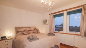 A bedroom with a bed, bedside table, lamp, window, and a view of the outside. - Roselea (Strath)