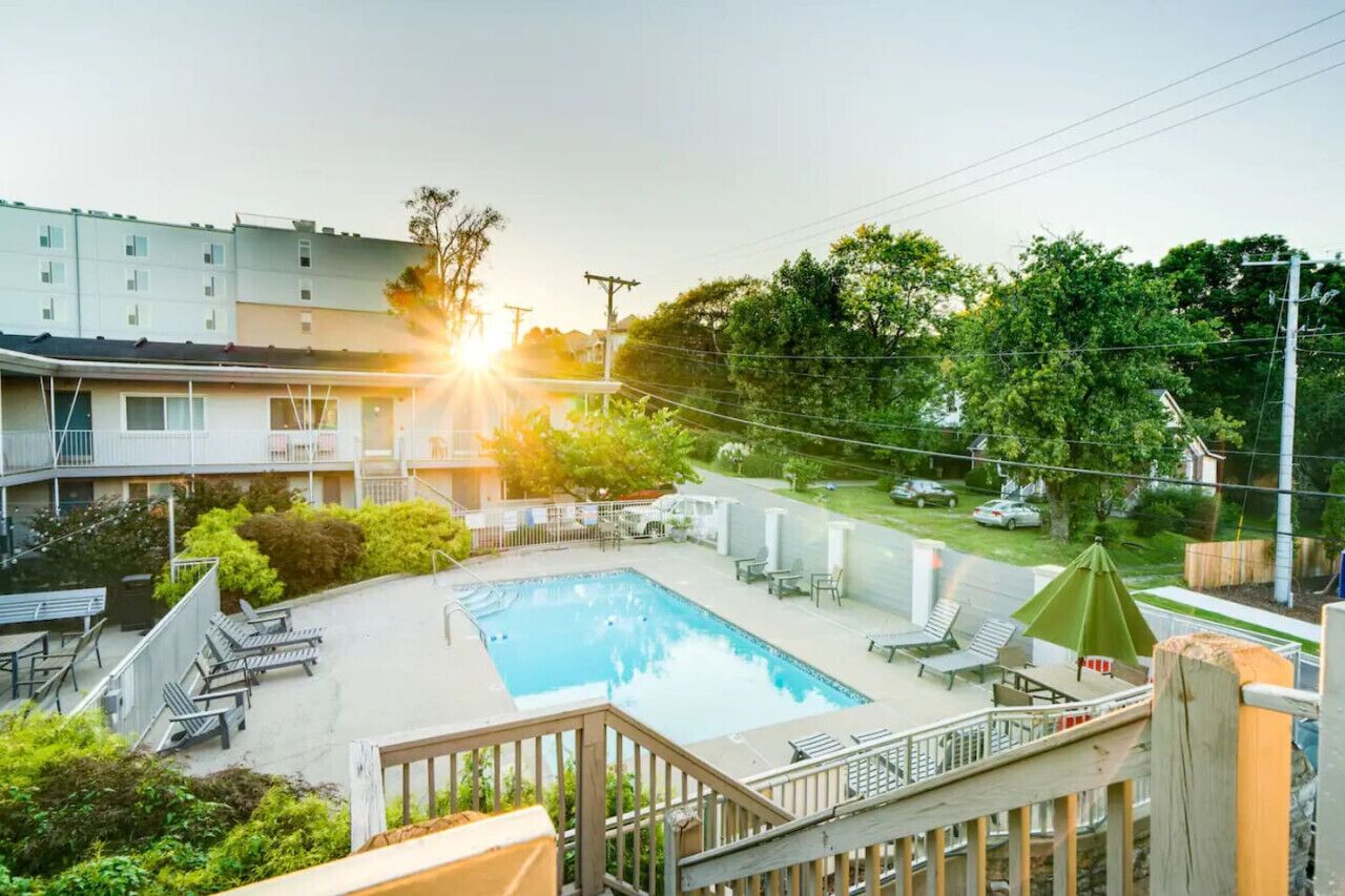 Piscina con sillas de descanso y sombrillas, rodeada de vegetación y un edificio de varios pisos.