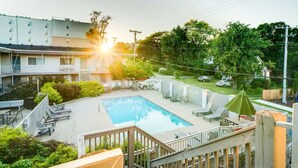 Piscina con sillas de descanso y sombrillas, rodeada de vegetación y un edificio de varios pisos.