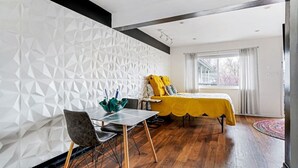Una habitación moderna con un papel tapiz de estampado geométrico, una cama amarilla, una mesa de comedor con sillas y un ventanal con cortinas.