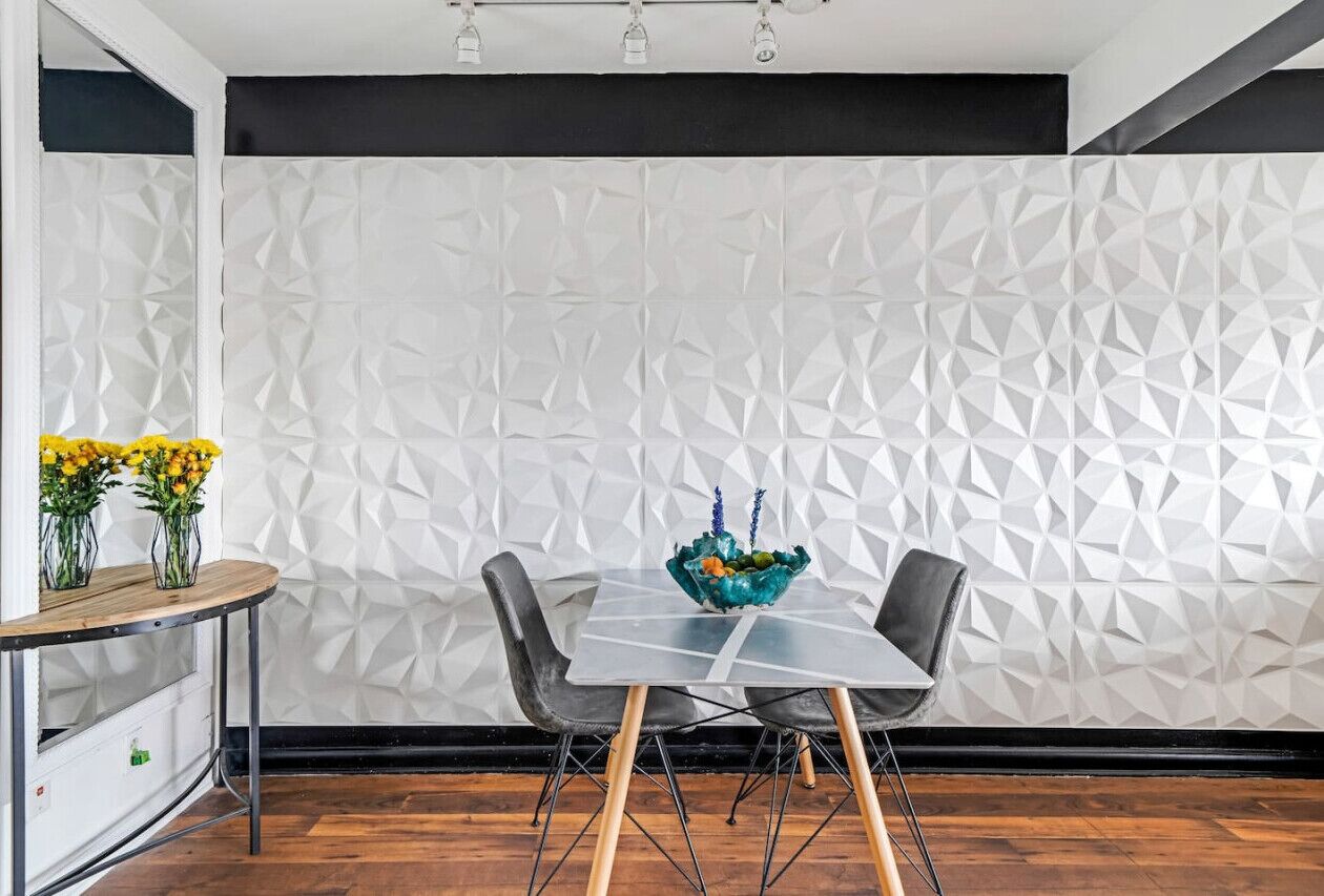 Un coin repas avec un mur orné d’un motif géométrique, une table à plateau en verre, deux chaises et un vase rempli de fleurs jaunes.