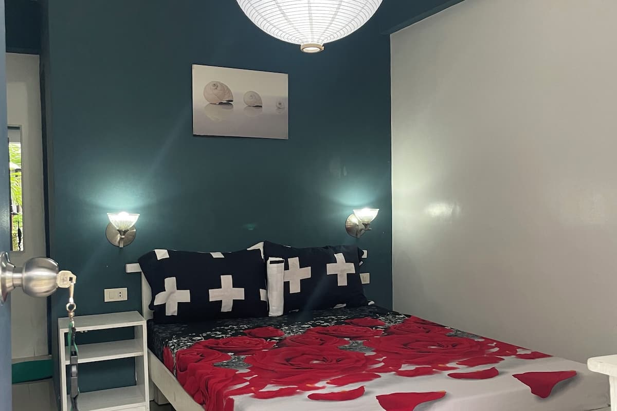 Un dormitorio con cama blanca, paredes verde azulado, una lámpara de techo blanca y una mesita de noche con lámpara.