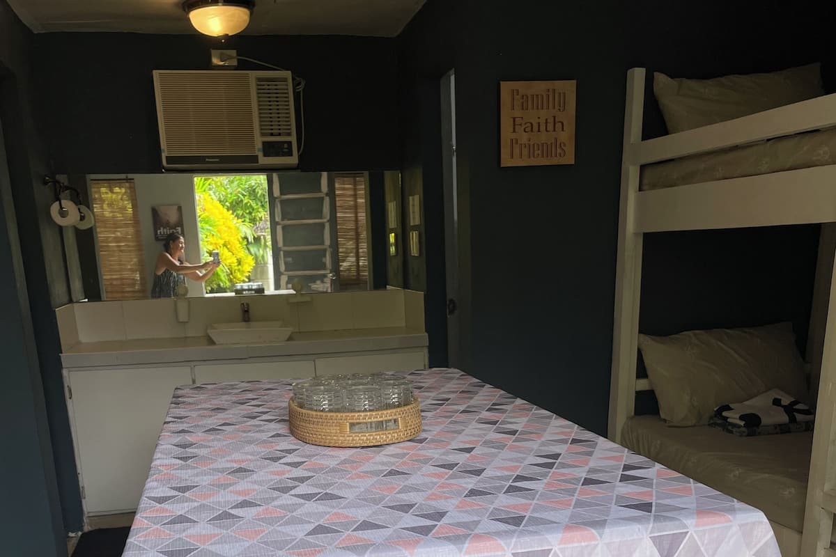 Una habitación con literas, una mesa central con mantel estampado, un equipo de aire acondicionado y un cartel que dice 'Family Faith Friends'.