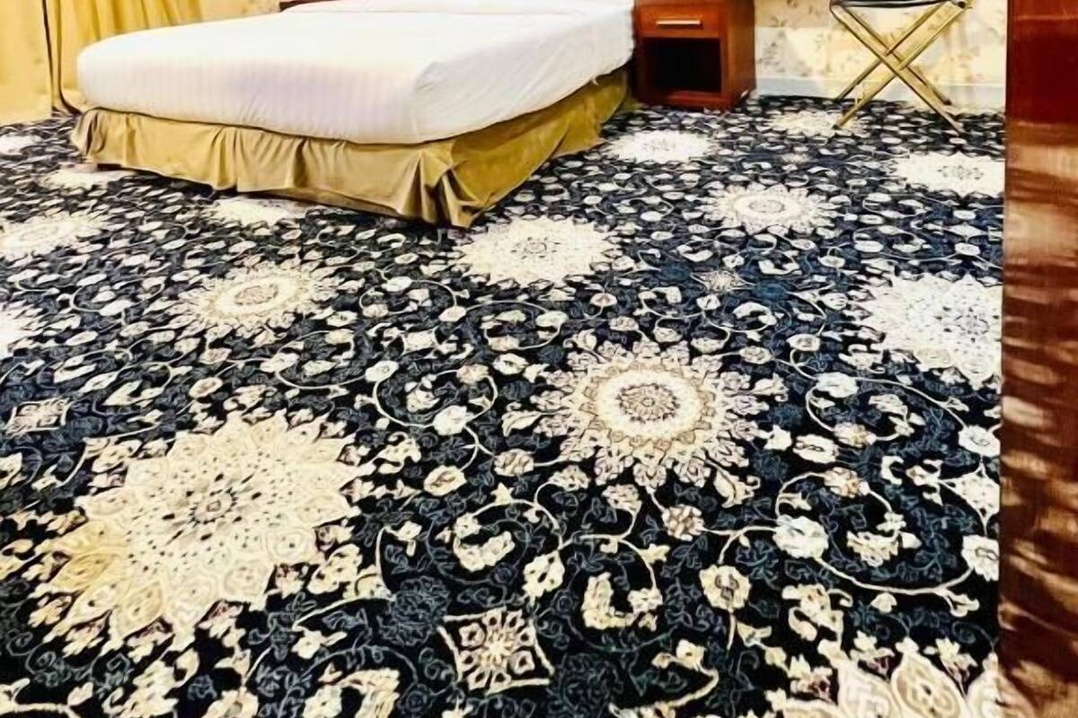 A hotel room with a large bed, a wooden headboard, a nightstand, and a patterned carpet.