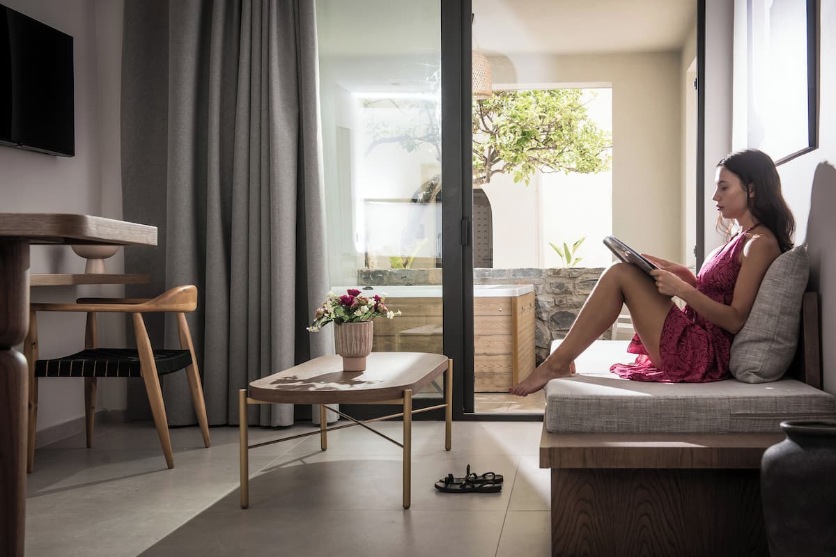 A modern living room with a sofa, a dining table with chairs, a small table with a vase of flowers, and a view of a garden through a sliding door.