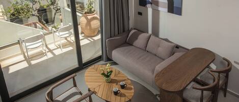 Una sala moderna con un sofá, una mesa de centro de madera y dos sillones. Hay un ventanal con vistas a un balcón y plantas en macetas.